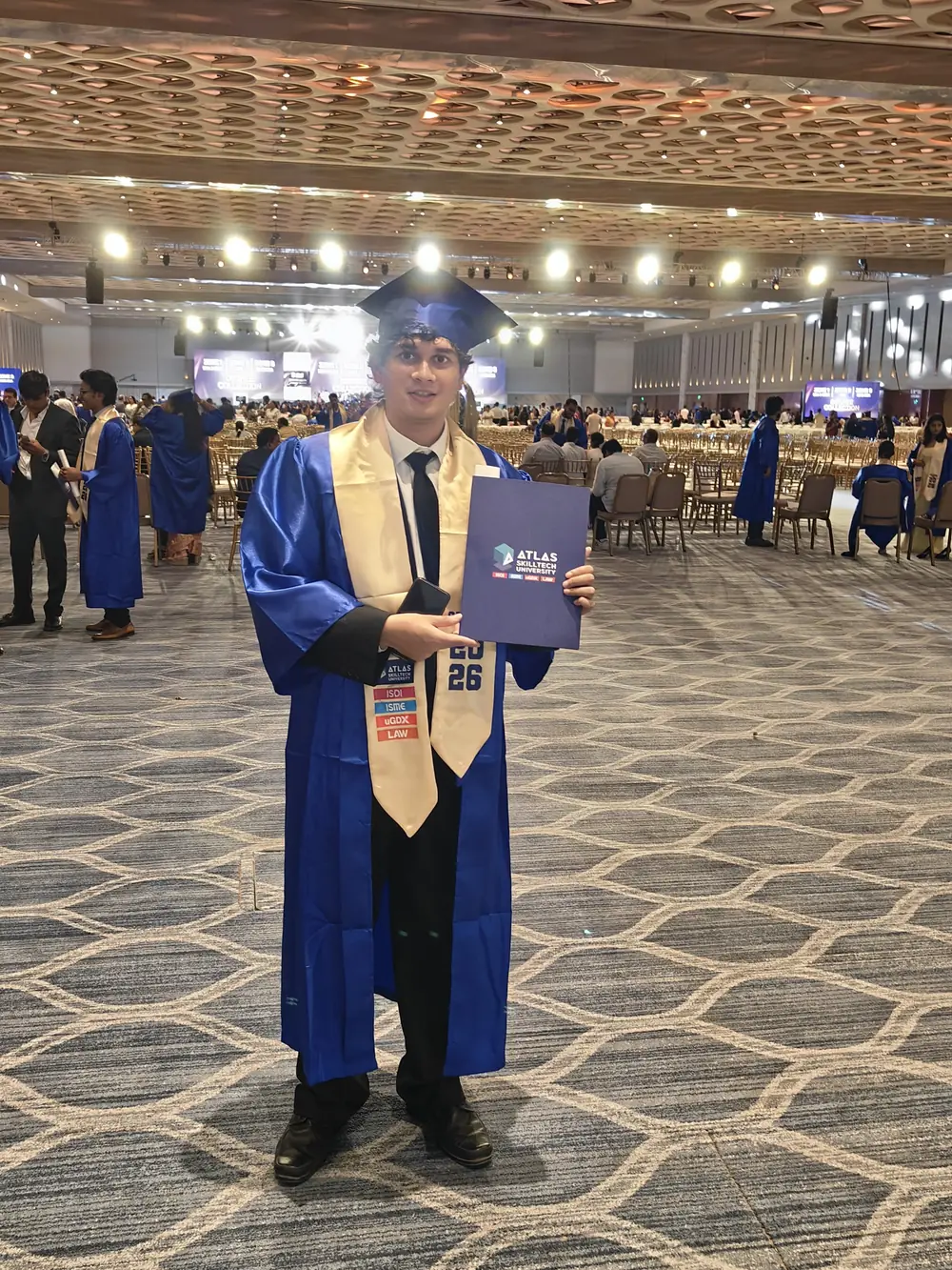 Keenan in a blue graduation gown holding an Atlas SkillTech University folder at the 2026 convocation